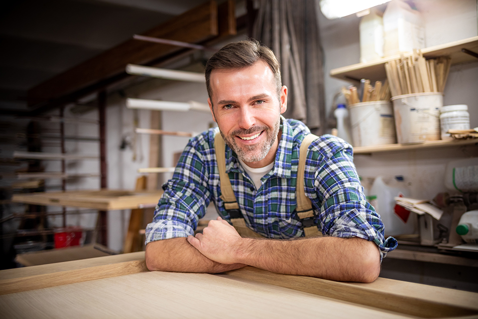 Proud, middle-aged male carpenter leaning on a work table in his workshop, smiling at the camera.