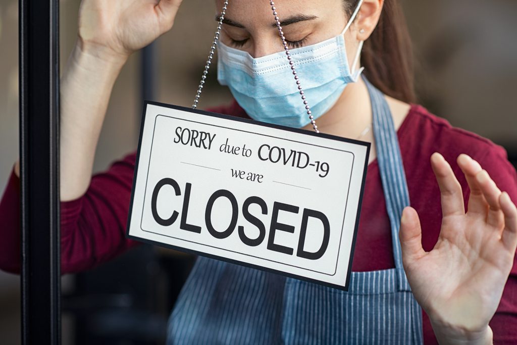 Stressed female owner standing behind sign in door window saying Sorry, close due to COVID-19.