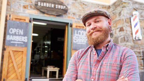 Hipster barber is smiling at camera, standing outside of his shoe doors.