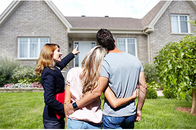 Female real estate agent next to young couple with arms entwined points to gray brick house in front of them.